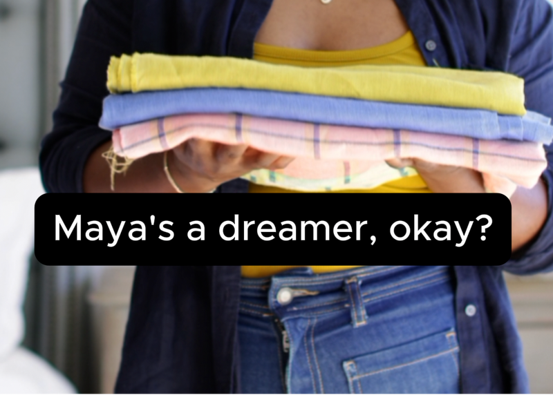 A woman with dark hair wearing a navy blue button-up shirt over a yellow top and blue jeans, holding a stack of folded colorful towels. A black text overlay at the bottom reads, 'Maya's a dreamer, okay?'.
