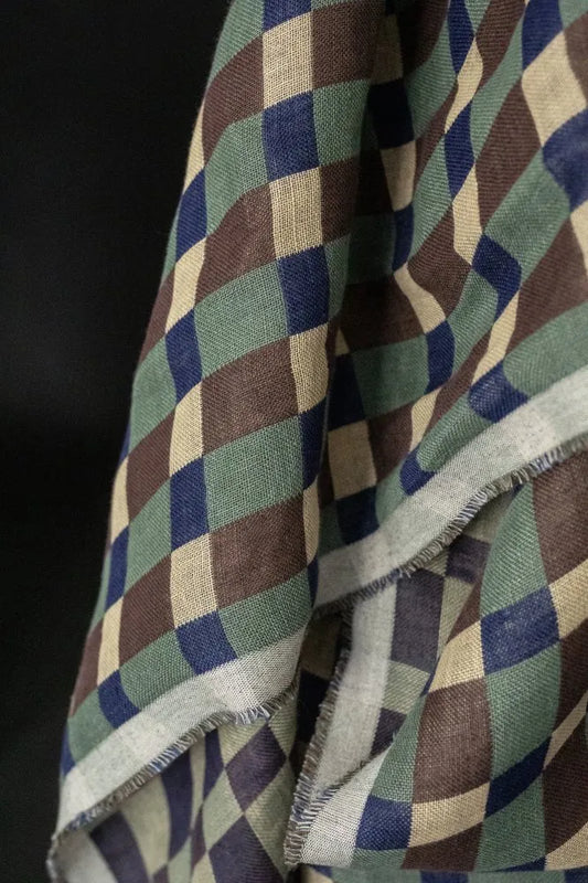 A close-up of a woven scarf or fabric with a geometric diamond pattern in olive green, navy blue, brown, and beige, featuring a frayed edge.